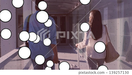 Walking couple rolling suitcases and carrying tote, crossbody bags in metal-framed glass corridor Walking couple rolling suitcases and carrying tote, crossbody bags in metal-framed glass corridor 139257130