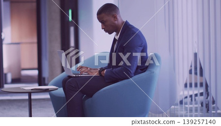 Typing man in business attire on lounge chair in corporate lounge, with laptop, sheet on table 139257140