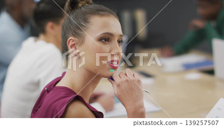 Listening woman in maroon blouse holding pen near chin in modern office, with documents, monitor Listening woman in maroon blouse holding pen near chin in modern office, with documents, monitor 139257507