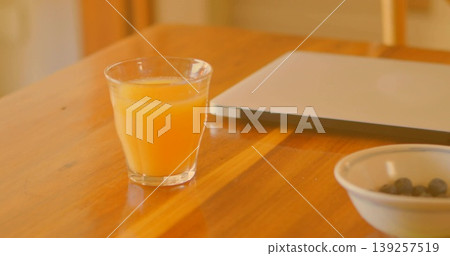 Displaying clear glass of orange juice sitting on dining table, with laptop and bowl of berries Displaying clear glass of orange juice sitting on dining table, with laptop and bowl of berries 139257519