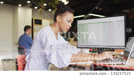 Typing woman in striped blouse leaning at laptop keyboard at desk, with monitor showing spreadsheet Typing woman in striped blouse leaning at laptop keyboard at desk, with monitor showing spreadsheet 139257525