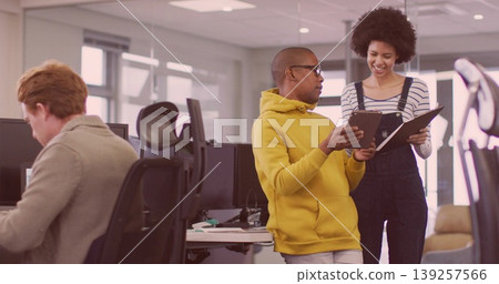 Standing man in yellow hoodie pointing at tablet in office, with coworker in overalls by monitors Standing man in yellow hoodie pointing at tablet in office, with coworker in overalls by monitors 139257566