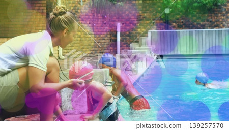 Teaching instructor kneeling at backyard pool guiding children wearing swim caps, holding strap Teaching instructor kneeling at backyard pool guiding children wearing swim caps, holding strap 139257570