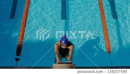 Leaning swimmer in blue cap and goggles, gripping starting block at outdoor pool, with orange ropes Leaning swimmer in blue cap and goggles, gripping starting block at outdoor pool, with orange ropes 139257590