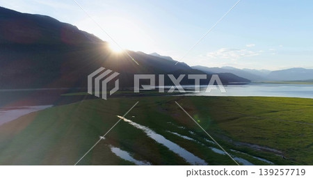 Shining central mountain ridge radiating sunbeams across marshy valley, with calm lake waters Shining central mountain ridge radiating sunbeams across marshy valley, with calm lake waters 139257719
