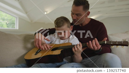 Father in maroon shirt guiding son playing acoustic guitar on sofa in living room, with focus Father in maroon shirt guiding son playing acoustic guitar on sofa in living room, with focus 139257740