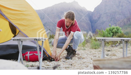 Crouching female camper securing yellow tent corner at rocky mountain campsite, with red duffel bag Crouching female camper securing yellow tent corner at rocky mountain campsite, with red duffel bag 139257786