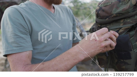 Inspecting bearded man wearing faded green V-neck T-shirt handling camouflage jacket in wooded area Inspecting bearded man wearing faded green V-neck T-shirt handling camouflage jacket in wooded area 139257799