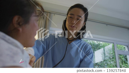 Listening Korean nurse in blue scrubs using stethoscope on patient chest in patient room, with bed Listening Korean nurse in blue scrubs using stethoscope on patient chest in patient room, with bed 139257848