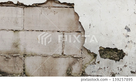A damaged wall where the white plaster is peeling and cracking, revealing the concrete block structure beneath. A damaged wall where the white plaster is peeling and cracking, revealing the concrete block structure beneath. 139257895
