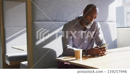 Holding smartphone, senior Black man wearing shirt sitting at modern lounge table with coffee cup Holding smartphone, senior Black man wearing shirt sitting at modern lounge table with coffee cup 139257946