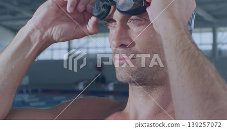 Shirtless athlete adjusting dark-tinted swim goggles in pool facility, water droplets on shoulders Shirtless athlete adjusting dark-tinted swim goggles in pool facility, water droplets on shoulders 139257972