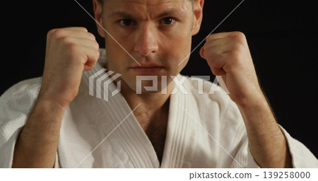Raising fists mid adult male holding defensive guard stance in studio, with white martial arts gi Raising fists mid adult male holding defensive guard stance in studio, with white martial arts gi 139258000