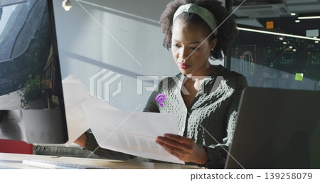Reviewing printed charts, woman in patterned blouse and headband sitting in office, with monitor Reviewing printed charts, woman in patterned blouse and headband sitting in office, with monitor 139258079