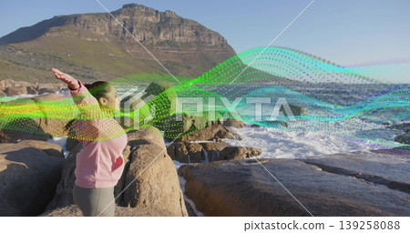 Standing Indian woman raising arms on coastal rocks, with ocean waves and digital ribbon overlay Standing Indian woman raising arms on coastal rocks, with ocean waves and digital ribbon overlay 139258088