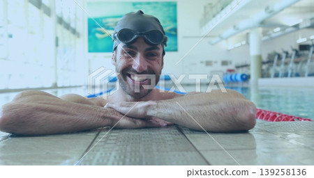 Smiling male swimmer resting on tiled pool deck with black swim cap, goggles and lane ropes Smiling male swimmer resting on tiled pool deck with black swim cap, goggles and lane ropes 139258136