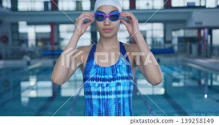 Adjusting purple goggles swimmer wearing blue swimsuit white swim cap at pool, with lane dividers 139258149