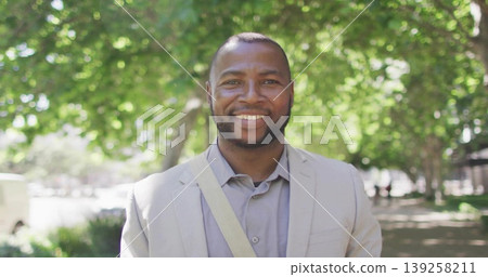 Standing man wearing light gray blazer on sunlit tree-lined walkway, with messenger bag strap Standing man wearing light gray blazer on sunlit tree-lined walkway, with messenger bag strap 139258211