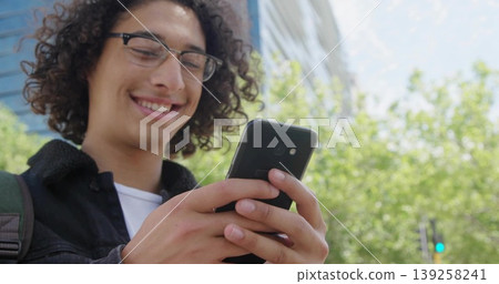 Grinning man wearing glasses and denim jacket checking smartphone on city street, carrying backpack Grinning man wearing glasses and denim jacket checking smartphone on city street, carrying backpack 139258241