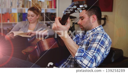 Strumming man playing acoustic guitar on armchair in living room, bookshelf and bicycle, copy space Strumming man playing acoustic guitar on armchair in living room, bookshelf and bicycle, copy space 139258289