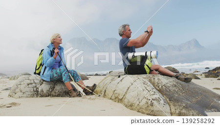 Sitting senior hiking couple capturing misty mountain view on coastal rocks, with backpacks Sitting senior hiking couple capturing misty mountain view on coastal rocks, with backpacks 139258292