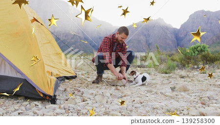 Squatting camper wearing red plaid shirt on rocky campsite, engaging terrier dog near yellow tent Squatting camper wearing red plaid shirt on rocky campsite, engaging terrier dog near yellow tent 139258300