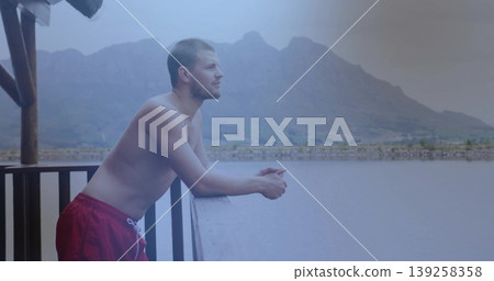 Leaning man wearing red trunks gazing toward mountain range over calm water at railing, copy space Leaning man wearing red trunks gazing toward mountain range over calm water at railing, copy space 139258358