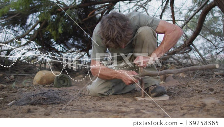 Kneeling man in green shirt, khaki pants pressing spindle into fireboard in scrubland, with tinder Kneeling man in green shirt, khaki pants pressing spindle into fireboard in scrubland, with tinder 139258365