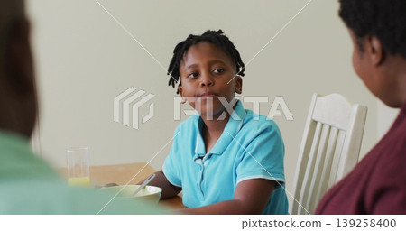 Black boy in polo eating cereal with spoon at dining table, with glass of orange juice Black boy in polo eating cereal with spoon at dining table, with glass of orange juice 139258400