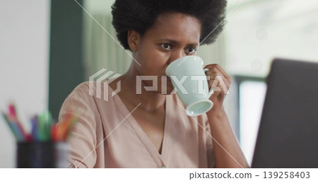 Holding light-colored ceramic mug, woman wearing pale pink blouse viewing laptop at home office Holding light-colored ceramic mug, woman wearing pale pink blouse viewing laptop at home office 139258403