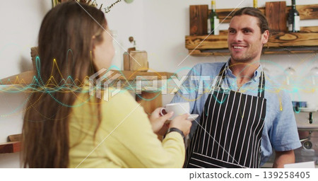 Serving barista wearing striped apron handing white coffee cup at shop counter, with coffee machine 139258405