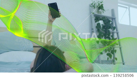 Scrolling smartphone, woman propping on pillow in bedroom with potted plant on shelf near window 139258506