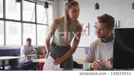 Leaning teenage female wearing sleeveless top pointing at printed charts in office, with monitor Leaning teenage female wearing sleeveless top pointing at printed charts in office, with monitor 139258553