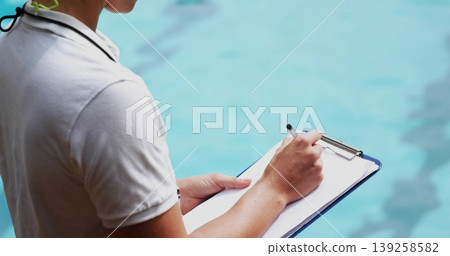 Lifeguard wearing white polo recording safety checks poolside, with clipboard pen and whistle cord Lifeguard wearing white polo recording safety checks poolside, with clipboard pen and whistle cord 139258582