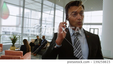 Standing Asian man in suit talking on smartphone in lobby, with coworkers using laptop, copy space Standing Asian man in suit talking on smartphone in lobby, with coworkers using laptop, copy space 139258615