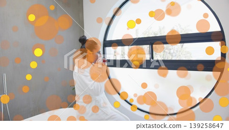 Holding white ceramic mug, Indian woman in bathrobe gazing through circular window in spa bathroom Holding white ceramic mug, Indian woman in bathrobe gazing through circular window in spa bathroom 139258647