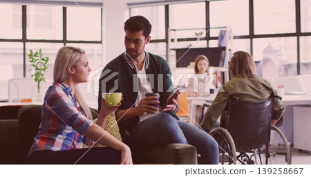 Talking blonde woman and coworker sitting on sofa in office lounge, holding green mug and tablet 139258667
