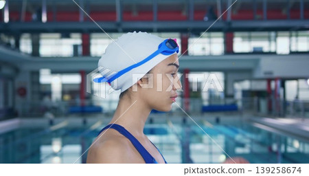 Standing female swimmer wearing swim cap and goggles on pool deck, with lane ropes, copy space Standing female swimmer wearing swim cap and goggles on pool deck, with lane ropes, copy space 139258674