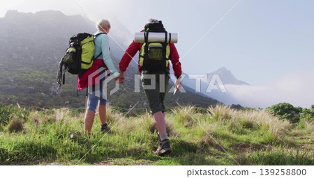 Walking mature couple ascending grassy mountain slope, carrying backpacks with rolled mats and map Walking mature couple ascending grassy mountain slope, carrying backpacks with rolled mats and map 139258800