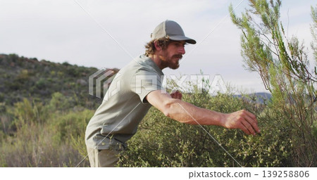 Leaning adult man examining medium-height bush in dry scrubland, with baseball cap and t-shirt Leaning adult man examining medium-height bush in dry scrubland, with baseball cap and t-shirt 139258806