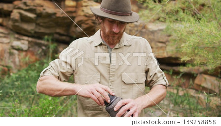 Twisting open container, bearded man wearing felt hat and khaki shirt outdoors at rock outcrop Twisting open container, bearded man wearing felt hat and khaki shirt outdoors at rock outcrop 139258858