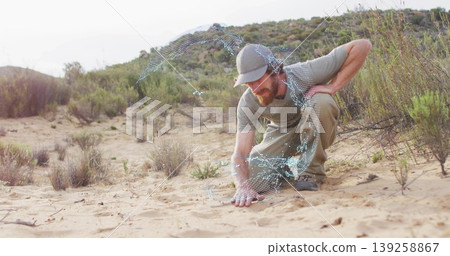 Explorer wearing baseball cap, over-ear headphones, touching sand in shrubland with digital swirl Explorer wearing baseball cap, over-ear headphones, touching sand in shrubland with digital swirl 139258867