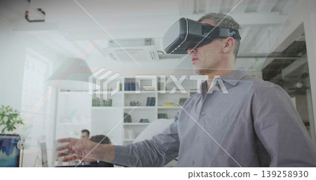 Standing mature White man wearing virtual reality headset reaching in open office, with computers Standing mature White man wearing virtual reality headset reaching in open office, with computers 139258930