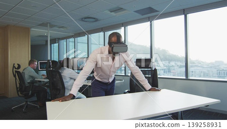 Leaning Black male wearing VR headset on white table in modern office, with dual monitors, mouse Leaning Black male wearing VR headset on white table in modern office, with dual monitors, mouse 139258931