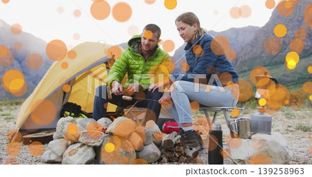 Roasting marshmallows couple in quilted jackets on mountain campsite with yellow tent fire ring Roasting marshmallows couple in quilted jackets on mountain campsite with yellow tent fire ring 139258963