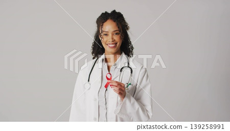 Smiling medical doctor wearing white coat and stethoscope, holding red ribbon at chest in studio Smiling medical doctor wearing white coat and stethoscope, holding red ribbon at chest in studio 139258991