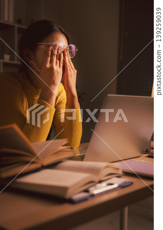 Young Asian woman in glasses rubbing tired eyes while using laptop in cozy home office at night, showing screen fatigue, study burnout, remote work stress, eyestrain and late productivity. Young Asian woman in glasses rubbing tired eyes while using laptop in cozy home office at night, showing screen fatigue, study burnout, remote work stress, eyestrain and late productivity. 139259039