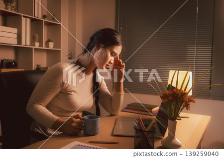 Young Asian woman working late on laptop in cozy home office, holding coffee and touching temple, conveying remote work stress, fatigue, concentration, deadline pressure and evening productivity. 139259040