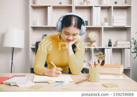 Young Asian woman student in headphones writing study notes at home desk with books, pencils and sticky notes. Focused learning, exam preparation, concentration, education and lifestyle concept. Young Asian woman student in headphones writing study notes at home desk with books, pencils and sticky notes. Focused learning, exam preparation, concentration, education and lifestyle concept. 139259066