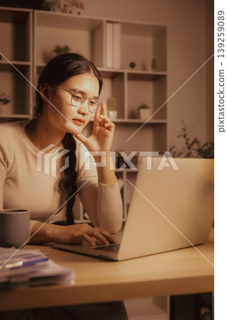 Young Asian woman in glasses working on laptop in cozy home office at night, showing deep focus, remote work, online study, problem solving, concentration and calm evening productivity. Young Asian woman in glasses working on laptop in cozy home office at night, showing deep focus, remote work, online study, problem solving, concentration and calm evening productivity. 139259089